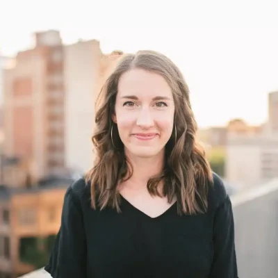 A young woman with wavy brown hair and hoop earrings stands outdoors, smiling gently. She wears a black top, and the background shows blurred city buildings in soft, natural light.