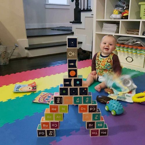 A smiling baby sits on a colorful foam mat next to a tall tower built from alphabet and symbol blocks. Toys and books are scattered around in a cozy, well-lit room.