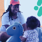 A woman in a coral cap smiles while playing with a toddler on blue rocking horse toys. The background has abstract shapes in light blue and tan colors.
