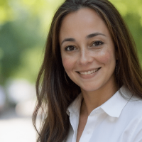 A woman with long brown hair, wearing a white collared shirt, smiles at the camera against a blurred outdoor background with green foliage.