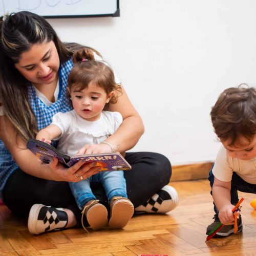 A woman sits on the floor reading a book to a young girl on her lap while a young boy plays with toys nearby on a wooden floor.