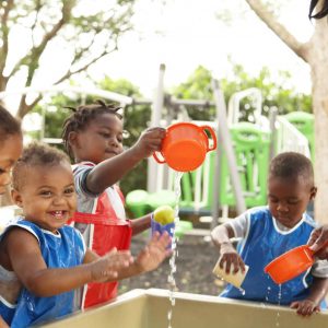 Four young children wearing blue and red aprons play with water and plastic cups outdoors, supervised by an adult. The children are smiling and laughing around a water table on a sunny day.
