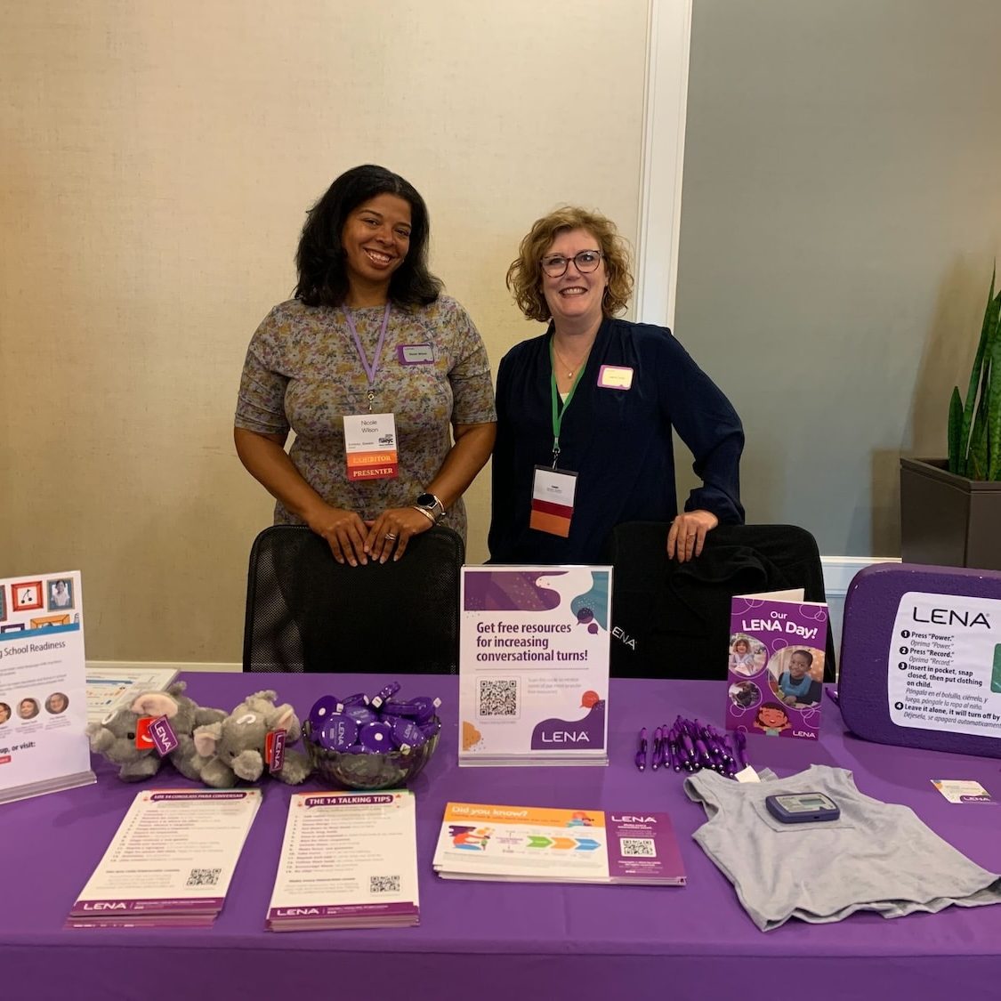 Two women stand behind a table covered with a purple cloth, displaying informational materials, flyers, and small giveaways at an indoor event or conference. A plant is visible in the background.