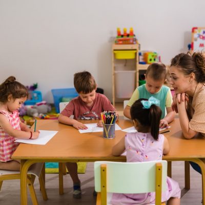Four young children sit around a table drawing with colored pencils while a woman, likely their teacher, attentively observes and smiles. The classroom is colorful and filled with toys and educational materials.