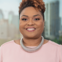 A woman with short curly hair smiles at the camera, wearing a light pink top and a large silver necklace, with a blurred cityscape in the background.