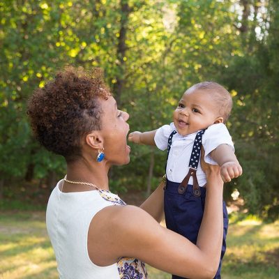 A woman joyfully lifts a smiling baby boy dressed in blue suspenders and a white shirt outdoors, with lush green trees in the background.