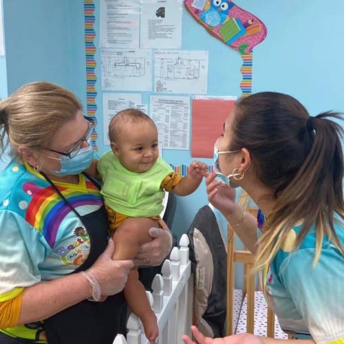 Two women wearing colorful uniforms and face masks interact with a smiling baby in a daycare setting. One woman holds the baby while the other gestures playfully. Bright decorations and charts are visible in the background.