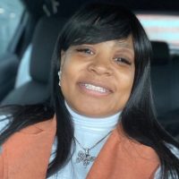A woman with straight black hair smiles while sitting in a car. She is wearing a white turtleneck, an orange-brown vest, and a butterfly pendant necklace.