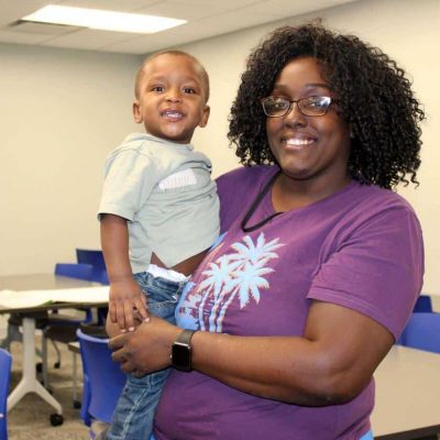 A smiling woman with curly hair, wearing glasses and a purple shirt, holds a happy young boy in a casual classroom setting with blue chairs and tables in the background.