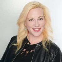 Smiling woman with long blonde hair wearing a black leather jacket and black top, posed against a plain light background.
