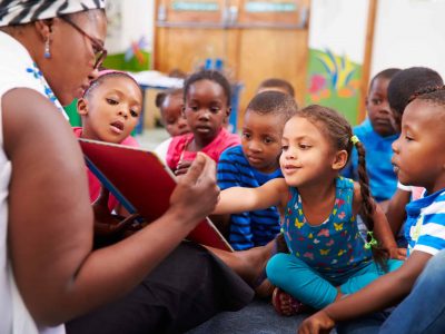 A group of young children sit on the floor, attentively listening to an adult reading a book to them. One girl reaches toward the book, while the other children watch and listen closely in a colorful classroom setting.