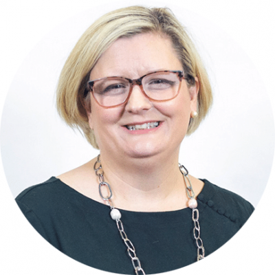 A woman with short blonde hair and glasses smiles at the camera. She is wearing a dark green top, a long necklace, and pearl earrings, with a white background behind her.