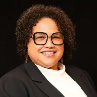 A smiling person with short curly hair, wearing large black glasses, gold earrings, a white top, and a black blazer, poses in front of a plain black background.