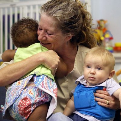 A smiling woman hugs a baby with curly hair while holding another baby with blond hair on her lap in a colorful, indoor setting.