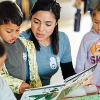 A woman reads a picture book to three young children gathered around a table. The children look at the book with interest, and all are wearing casual clothes. The setting appears to be a classroom or daycare.